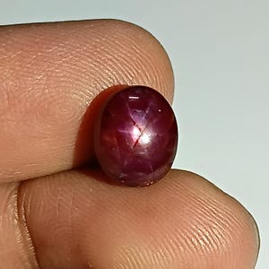 Könnte beinhalten: Ein polierter, ovaler Rubin-Edelstein. Der Edelstein hat eine tiefrote Farbe mit einem sternartigen Effekt auf seiner Oberfläche. Der Stein wird zwischen Fingern gehalten, um seine Größe und Form zu zeigen. Der Rubin ist ein natürlicher Edelstein.