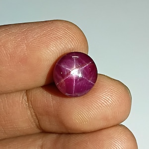 Könnte beinhalten: Ein runder, polierter Sternrubin-Edelstein. Der Edelstein hat eine tiefe, satte rote Farbe mit einem sechsstrahligen Sternmuster, das über seine Oberfläche sichtbar ist. Der Edelstein wird in einer Hand gehalten, um seine Größe und seinen Glanz zu zeigen.