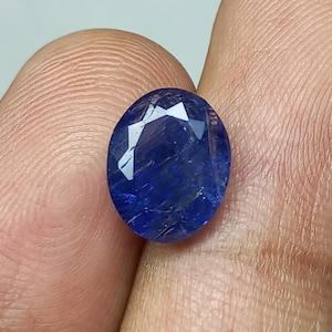 May include: Oval-cut, deep blue gemstone with facets reflecting light. The gem has a polished surface and a rich, saturated color. The stone is held between fingers, showcasing its size and clarity. A beautiful piece for jewelry.