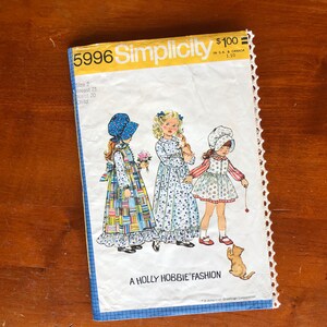 Op de afbeelding: Een vintage Simplicity naaipatroon voor kinderjurken. Het patroon is voor de maten 2, 21 en 20. Het patroon toont drie meisjes die verschillende jurken en hoeden dragen. Het patroon is getiteld "A Holly Hobbie Fashion".
