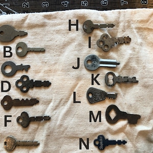 Könnte beinhalten: Eine Sammlung von 14 Vintage-Schlüsseln, die mit den Buchstaben A bis N beschriftet sind, auf einer weißen Stoffoberfläche angeordnet. Die Schlüssel sind aus Metall und haben verschiedene Formen und Größen.