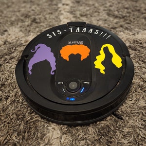 Puede incluir: Un robot aspirador Shark negro con una pegatina morada, naranja y amarilla de tres brujas con el texto "Sis-taaaas!!" en la parte superior.
