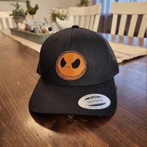 Könnte beinhalten: Schwarze und orangefarbene Baseballkappe mit einem gestickten Lederpatch, der das Gesicht von Jack Skellington aus dem Film Nightmare Before Christmas zeigt. Die Kappe hat ein Netzrücken und ein schwarzes Etikett "The Authentic Yupoong Snapback".