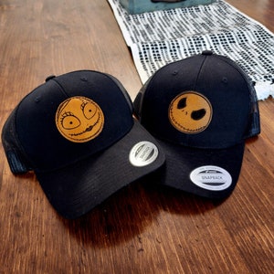 Könnte beinhalten: Zwei schwarze Mesh-Trucker-Caps mit Leder-Patches, die die Gesichter von Jack Skellington und Sally aus dem Film Nightmare Before Christmas zeigen. Die Mützen haben den Text "The Authentic Snapback" auf der Rückseite.