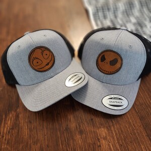 Könnte beinhalten: Zwei graue und schwarze Trucker-Caps mit Leder-Patches. Ein Patch zeigt ein zusammengenähtes lächelndes Gesicht, der andere ein zusammengenähtes missmutiges Gesicht. Die Caps haben ein schwarzes Netzrücken und ein silbernes Etikett mit der Aufschrift "The Authentic Snapback" und einem Logo.