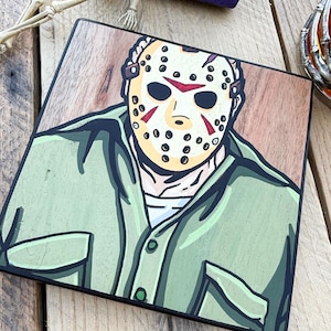 May include: A square wooden plaque featuring a hand-painted illustration of a character wearing a cream-colored hockey mask with red markings and a green button-down shirt. The artwork is rendered in a bold, graphic style with thick black outlines.