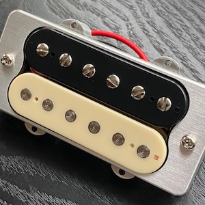 May include: A guitar pickup with a brushed metal base. It has two rectangular pickups, one black and one cream-coloured, each with visible screws. Red wiring is visible.
