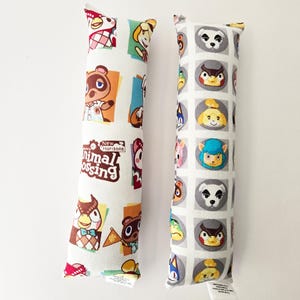 May include: Two rectangular fabric pillows featuring Animal Crossing New Horizons character prints. The left pillow displays various characters in colorful squares with the game logo. The right pillow features a grid of circular character portraits.
