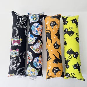 May include: Four cat toys with colorful patterns. One is black with white cat skeleton and sugar skull designs. Another is black with sugar skull cat faces. The third is orange with black cat silhouettes. The last is yellow with black cat silhouettes.