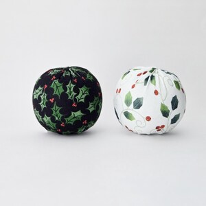 Può includere: Due ornamenti rotondi in tessuto. Uno è nero con foglie di agrifoglio verdi e bacche rosse. L'altro è bianco con foglie verdi e bacche rosse. Decorazioni festive natalizie.