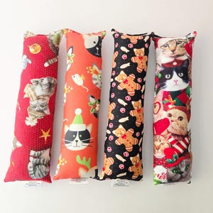 May include: Four rectangular fabric cat toys featuring festive holiday prints. The first is red with kittens, the second is orange with cat faces and mice, the third is black with gingerbread cookies, and the fourth features a collage of cats.