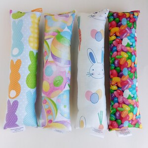 May include: Four Easter-themed fabric pillows with different designs. The pillows are rectangular and have a white background. The first pillow has a pattern of purple and orange bunnies. The second pillow has a pattern of colorful Easter eggs. The third pillow has a pattern of a white bunny with a carrot. The fourth pillow has a pattern of colorful jelly beans.
