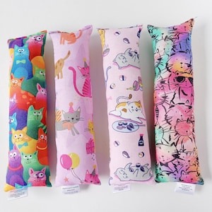 May include: Four catnip toys featuring different cat designs. The toys are long and rectangular, with one featuring a rainbow pattern, one with a sushi pattern, one with a birthday party pattern, and one with a colourful cat pattern.