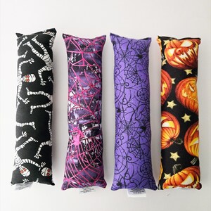 May include: Four rectangular fabric catnip kicker toys arranged vertically. The first features white mummies on black, the second has pink spiderwebs on purple, the third shows black spiderwebs on purple, and the fourth displays orange jack-o'-lanterns on black.
