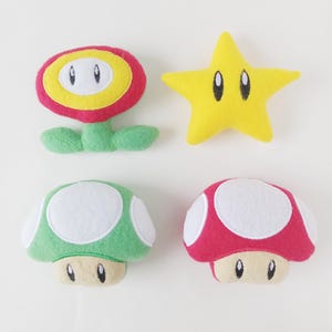 May include: Four plush toys from the Super Mario Bros. video game series. The toys are a red flower with yellow petals and a white face, a yellow star with a white face, a green mushroom with a white top and a white face, and a red mushroom with a white top and a white face.