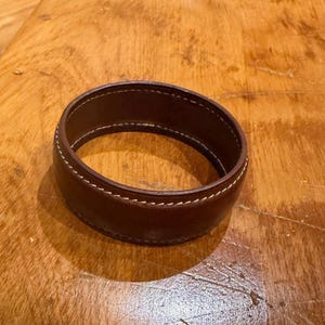 May include: A brown leather cuff bracelet with white stitching. The bracelet is circular and appears to be made of a single piece of leather. The stitching is visible along the edges of the bracelet.