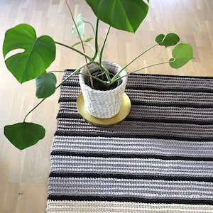 Puede incluir: Una alfombra de crochet a rayas en tonos beige, gris y negro. La alfombra está sobre un suelo de madera y una planta en maceta está en primer plano.