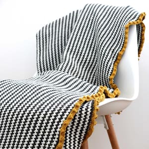 Può includere: Una coperta a righe bianche e nere in crochet con un bordo in crochet giallo. La coperta è drappeggiata su una sedia bianca con gambe in legno.