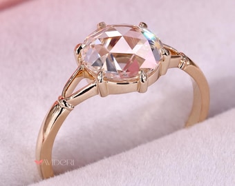 May include: A gold engagement ring with a large, oval-cut diamond. The ring is set in a delicate, prong setting and is displayed in a pink ring box. The band has a simple, elegant design.