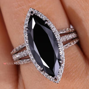 Può includere: Anello con pietra nera a taglio marquise, circondata da piccole gemme chiare. L'anello ha una doppia fascia con ulteriori piccole gemme. L'anello è incastonato in metallo color argento.