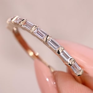 May include: A gold ring with a band set with a line of rectangular, clear gemstones. The ring is held against a soft pink background, highlighting the sparkle of the stones. The design is simple and elegant.
