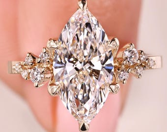 Könnte beinhalten: Ein Verlobungsring mit Diamanten im Marquiseschliff und einem goldenen Band. Der Ring hat einen großen, zentralen Diamanten und kleinere Akzentdiamanten, die auf beiden Seiten angeordnet sind und ein blütenähnliches Design bilden. Der Ring wird von den Fingern einer Person gehalten.