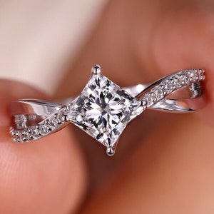 Può includere: Un anello di fidanzamento in argento con un diamante di forma quadrata e una fascia intrecciata decorata con diamanti più piccoli.