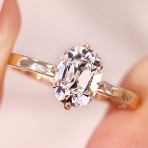 May include: A gold ring with a large, oval-cut diamond. The diamond is clear and multifaceted, reflecting light. The ring band is a simple, polished gold. The ring is held up by a person's fingers.