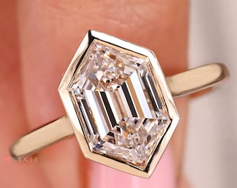 May include: A gold ring featuring a large, clear, hexagonal-cut diamond. The diamond is set in a bezel setting, and the band is a simple, polished gold. The ring is held against a blurred pink background.