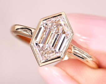 May include: A gold ring featuring a large, clear, hexagonal-cut gemstone. The gemstone is set in a bezel setting, and the band is a simple, polished gold. The ring is held against a soft pink background.