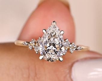 May include: A pear-shaped diamond engagement ring with a gold band and smaller diamonds set on the band. The ring is on a finger.