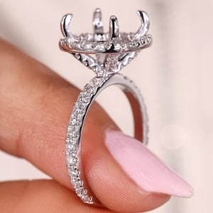 May include: A silver engagement ring setting with a halo design. The ring features a band encrusted with small, sparkling stones. The top of the ring has prongs to hold a center stone. The ring is held between two fingers.