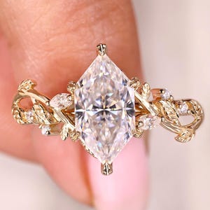 May include: A gold engagement ring with a large, marquise-cut diamond as the focal point. The band is decorated with intricate leaf and vine patterns, and small accent diamonds. The ring is set against a soft pink backdrop.