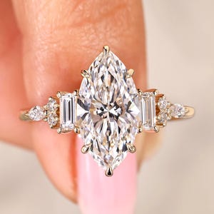 May include: A gold ring featuring a large, marquise-cut diamond as the centerpiece, flanked by two rectangular diamonds and smaller accent stones. The ring's band is a delicate gold color.