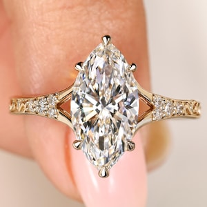 May include: A gold ring featuring a large marquise-cut center stone held by six prongs. The band has a split shank design adorned with small round accent stones and intricate filigree metalwork along the sides of the setting.