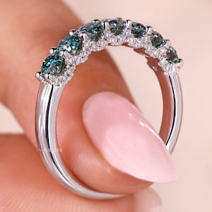 Peut inclure: Une bague en argent ornée d'une rangée de pierres précieuses de couleur teal, chacune entourée de petites pierres claires. La bague est présentée, mettant en valeur son design et l'éclat des gemmes. L'anneau de la bague est lisse et poli.