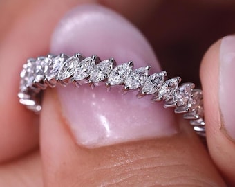May include: A silver ring with a continuous row of marquise-cut diamonds. The ring is held between fingers with manicured nails. The diamonds are clear and sparkle, reflecting light. The ring is a piece of jewelry.