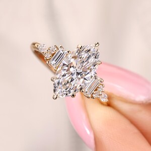 May include: A gold engagement ring featuring a large, marquise-cut diamond as the centerpiece. The ring is accented with smaller, baguette-cut and round diamonds on the band and surrounding the main stone. The ring is held up by a person with pink nail polish.