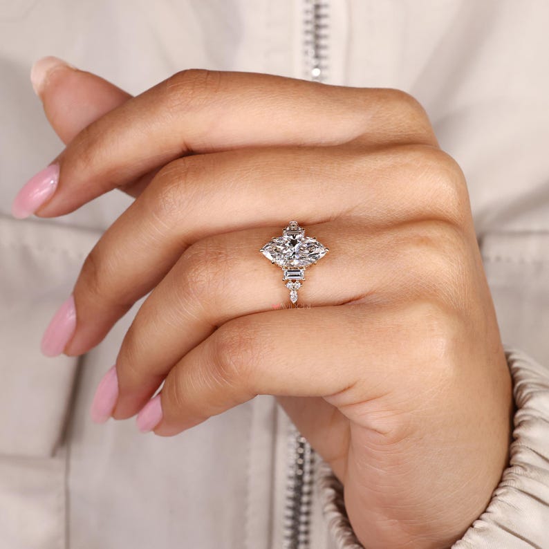 May include: A diamond ring featuring a marquise-cut center stone and smaller accent stones. The ring has a unique geometric design with a delicate band. The ring is set on a hand with pink nail polish.