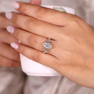 K&ouml;nnte beinhalten: Ein Verlobungsring mit einem Diamanten im Marquiseschliff. Das Design des Rings umfasst einen zentralen, l&auml;nglichen Diamanten, erg&auml;nzt durch kleinere Steine und detaillierte Metallarbeiten. Das Band zeigt ein Blumen- oder Rankenmuster.