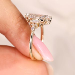 May include: A gold ring featuring a large, pear-shaped gemstone as the centerpiece, flanked by smaller, rectangular and round accent stones. The ring's band is a simple, polished gold. The ring is held up by a person's fingers.