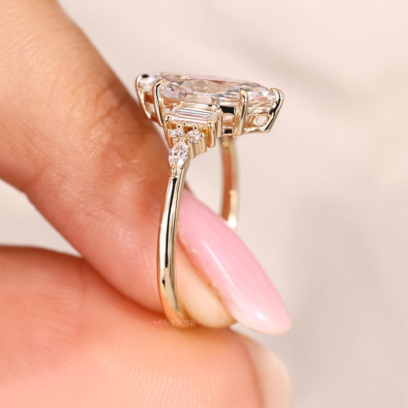 May include: A gold ring featuring a large, pear-shaped gemstone as the centerpiece, flanked by smaller, rectangular and round accent stones. The ring's band is a simple, polished gold. The ring is held up by a person's fingers.