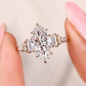 May include: A gold ring featuring a large, marquise-cut diamond as the centerpiece, flanked by two rectangular diamonds and smaller accent stones. The ring's band is also gold, creating a cohesive and elegant design.