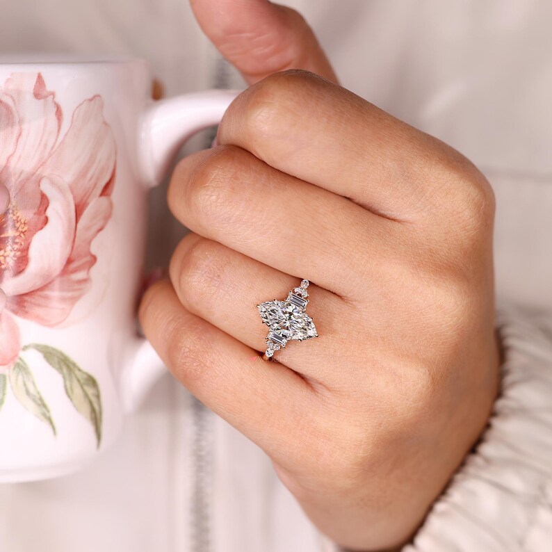 May include: A marquise-cut diamond engagement ring with a cluster of smaller diamonds on each side. The ring is set in a white metal band and sparkles brightly. The ring is on a person's finger.