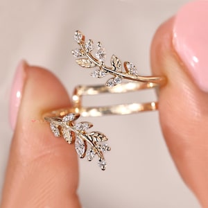 Può includere: Un anello color oro con un design a foglia, adornato con piccole e scintillanti pietre preziose trasparenti. L'anello si avvolge attorno al dito, con due elementi a forma di foglia. Le pietre preziose sono incastonate nelle foglie.