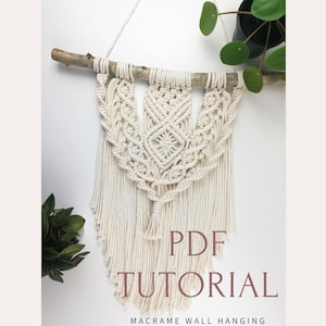 Könnte beinhalten: Ein weißer Makramee-Wandbehang mit einem Ast oben und einem PDF-Tutorial zur Herstellung. Der Text "PDF TUTORIAL" ist in braunen Buchstaben unter dem Behang geschrieben. Der Text "MACRAME WALL HANGING" ist in kleineren braunen Buchstaben unter dem Tutorial-Text geschrieben.