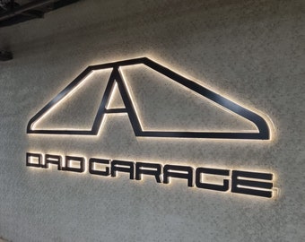 Könnte beinhalten: Ein schwarz-weißes Schild mit dem Text "DAD GARAGE" und einem stilisierten Buchstaben "A" mit leuchtender Umrandung.