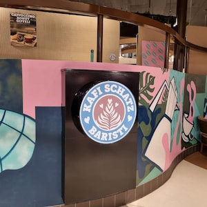 May include: A black sign with a blue circle and white text that reads "Kaffi Schatz Baristi" with a stylized leaf design inside the circle. The sign is mounted on a wall with pink, blue, and green painted designs.