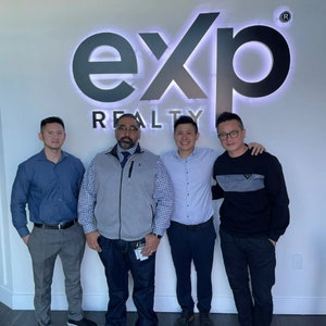 May include: Four men stand in front of a white wall with a black and white sign that reads "exp REALTY" with a registered trademark symbol. The men are wearing casual clothing.