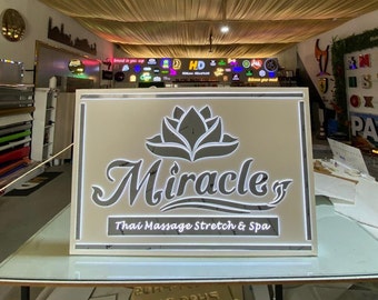 May include: A white sign with a silver lotus flower and the text "Miracle Thai Massage Stretch & Spa". The sign is illuminated with white lights.